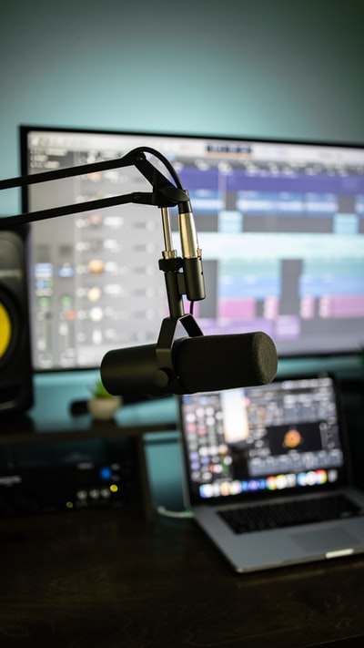 Close-up of a microphone on a boom arm in front of computer screens showing audio software, dark moody two-tone tones